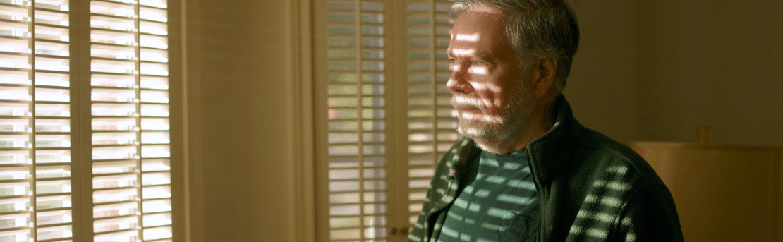 Worried man looking out through the blinds in his living room.